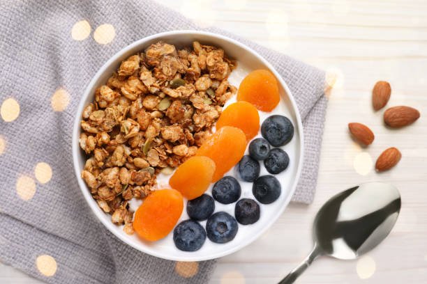  A bowl of granola topped with blueberries, almonds, and apricot dry fruit, placed on a wooden table.