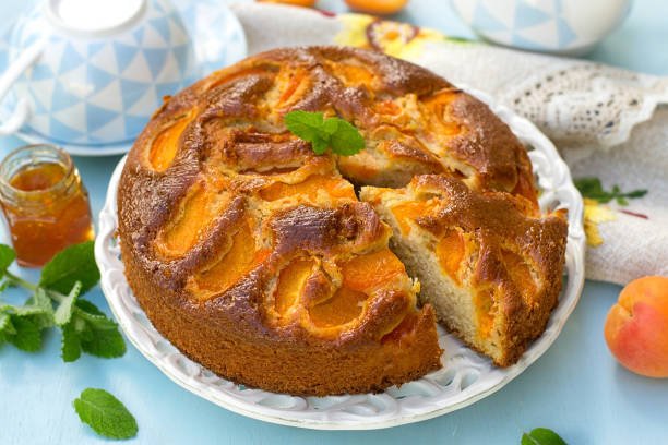 A beautifully arranged cake topped with fresh peaches, served on an elegant plate alongside apricot dry fruit pieces.