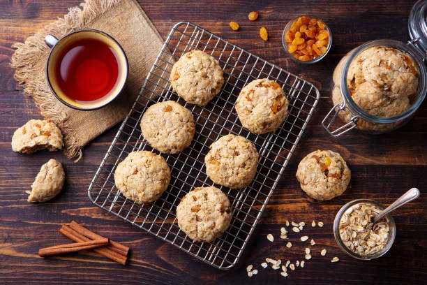 A cooling rack displays freshly baked cookies alongside a cup of tea and apricot dry fruits, creating a cozy snack scene.