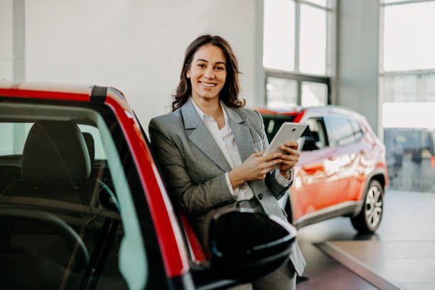 A professional woman in a suit holds a tablet, standing confidently in front of a car, symbolizing automobile insurance.